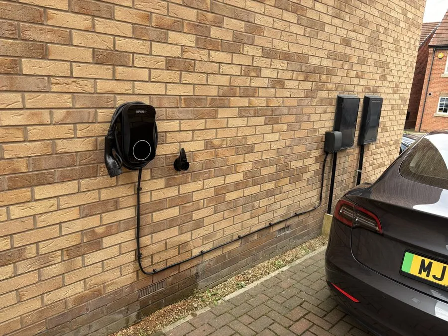 Black home EV charger on a brick wall beside a Tesla on a driveway