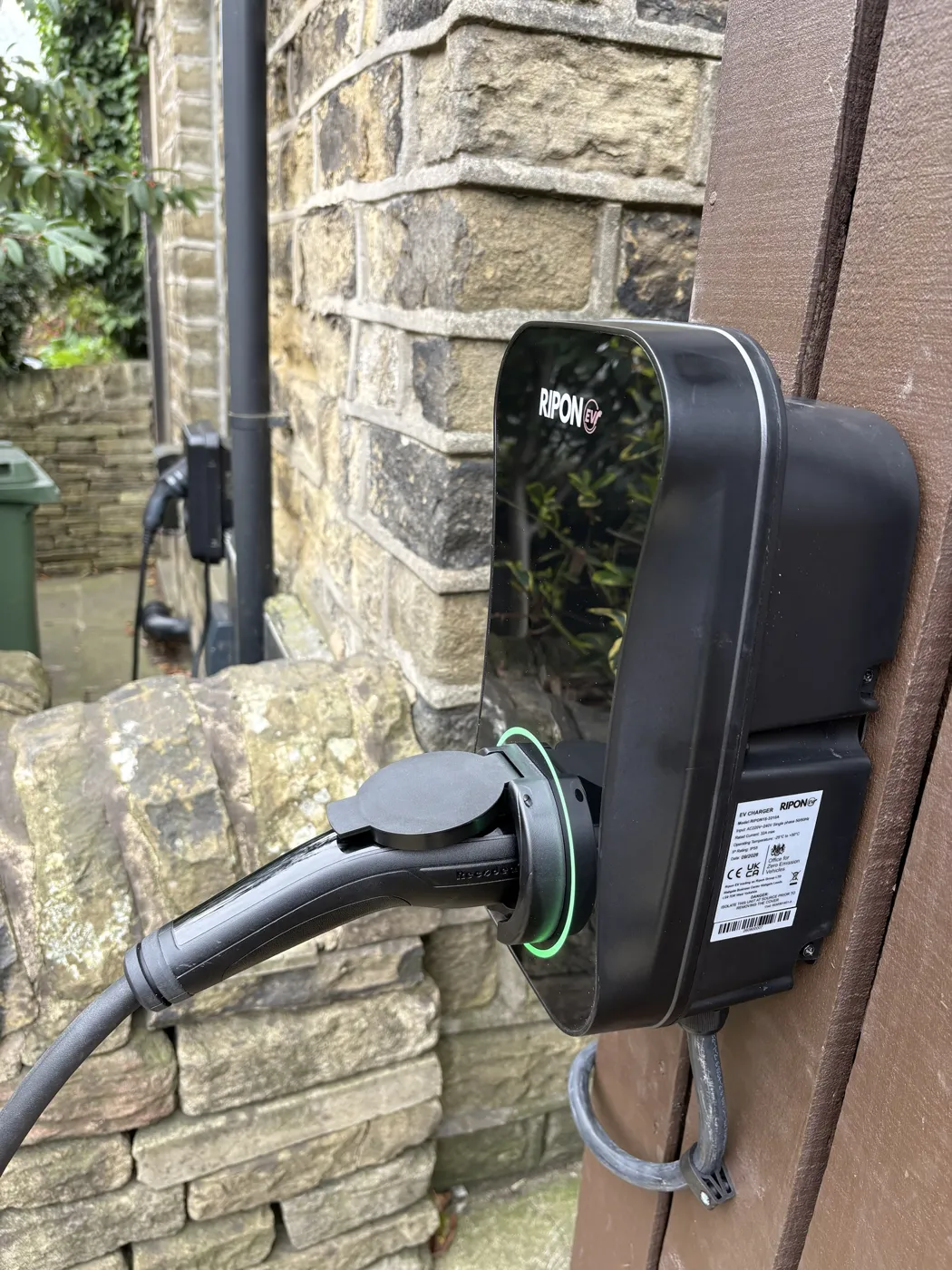 RiponEV charger on a timber post beside a stone house, plugged in with the green ring lit