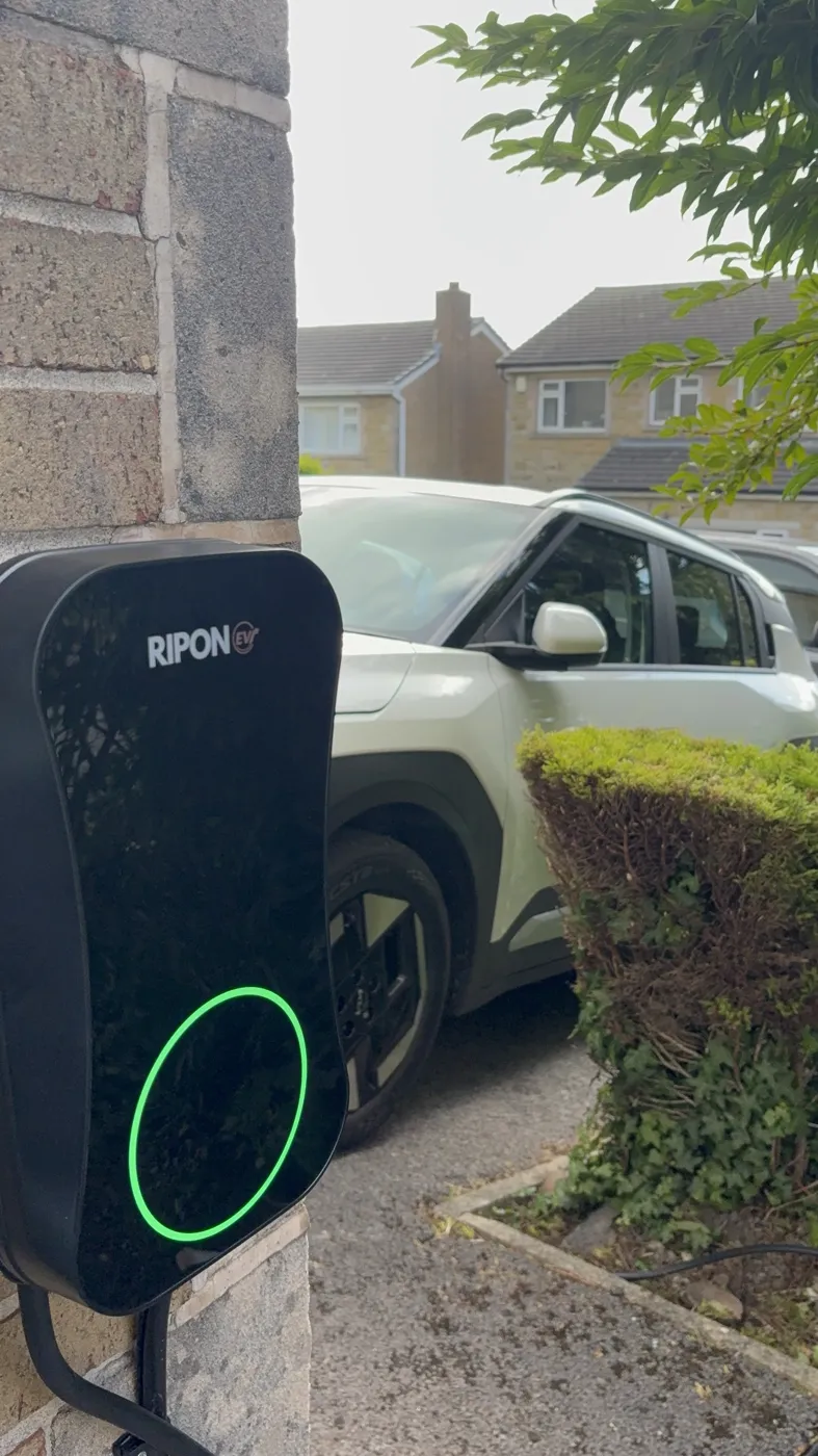 RiponEV charger glowing green on a stone wall with the car on the drive behind