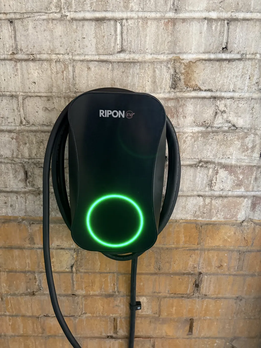 Tethered RiponEV charger with green status ring and cable wrapped on a brick wall