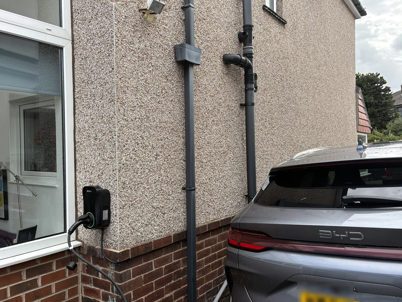 Wide view of a RiponEV charger on the brick plinth of a pebbledashed house beside the drive